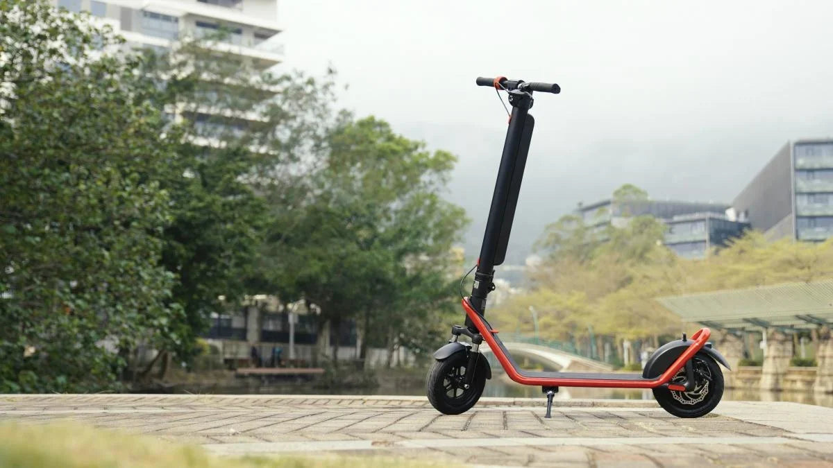 Schwarzer HX E-Scooter mit rotem Rahmen auf dem Ständer, gepflasterte Promenade mit Bäumen und modernen Gebäuden