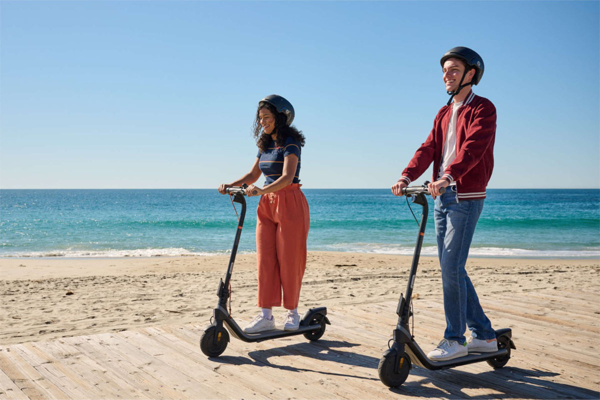 Zwei Personen mit Helm auf je einem E-Scooter auf einem Holzsteg am Strand – kann so einer das Auto zur Arbeit ersetzen?