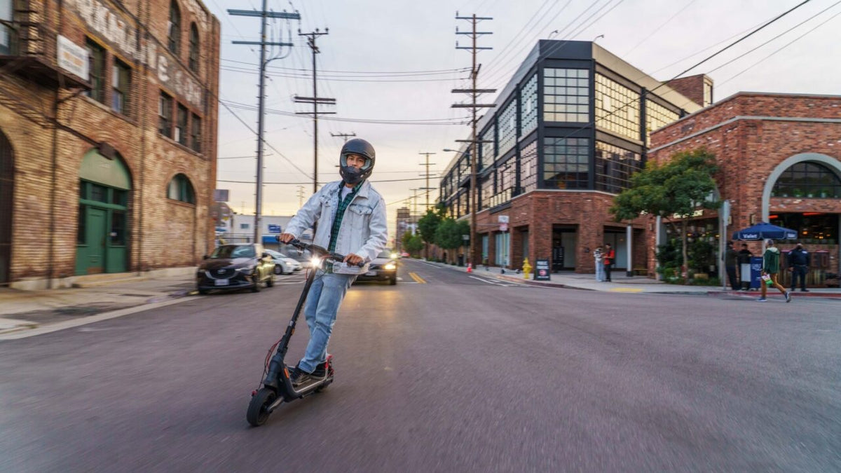 Mann mit schwarzem Integralhelm fährt E-Scooter mit eingeschaltetem Licht durch eine Stadtstraße in der Dämmerung