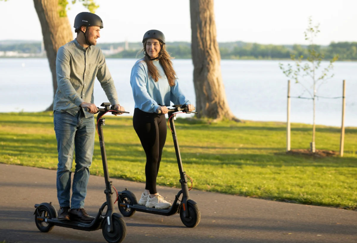 Zwei Personen mit Helm fahren NAVEE E-Scooter auf einem asphaltierten Weg an einem See