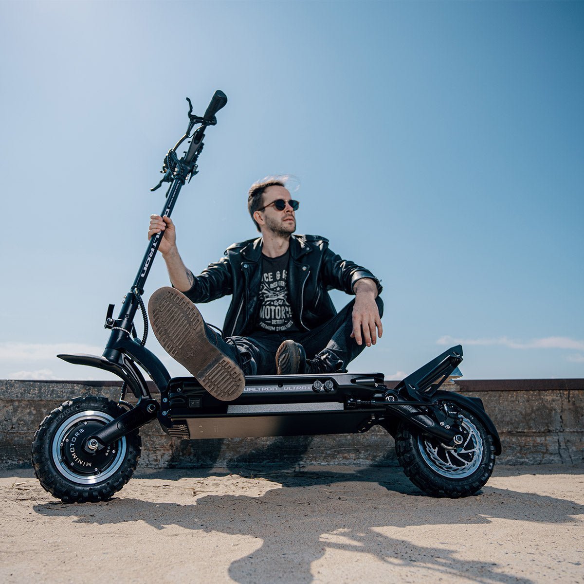 Mann in Lederjacke und Sonnenbrille sitzt auf einem schwarzen Dualtron Ultra mit groben Geländereifen auf Sand