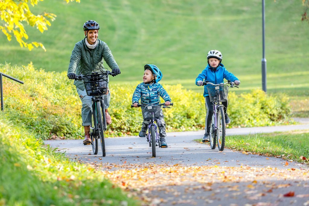Kvinna med cykelhjälm cyklar med två barn längs en parkväg en höstdag – alla tre cyklarna har korg på styret