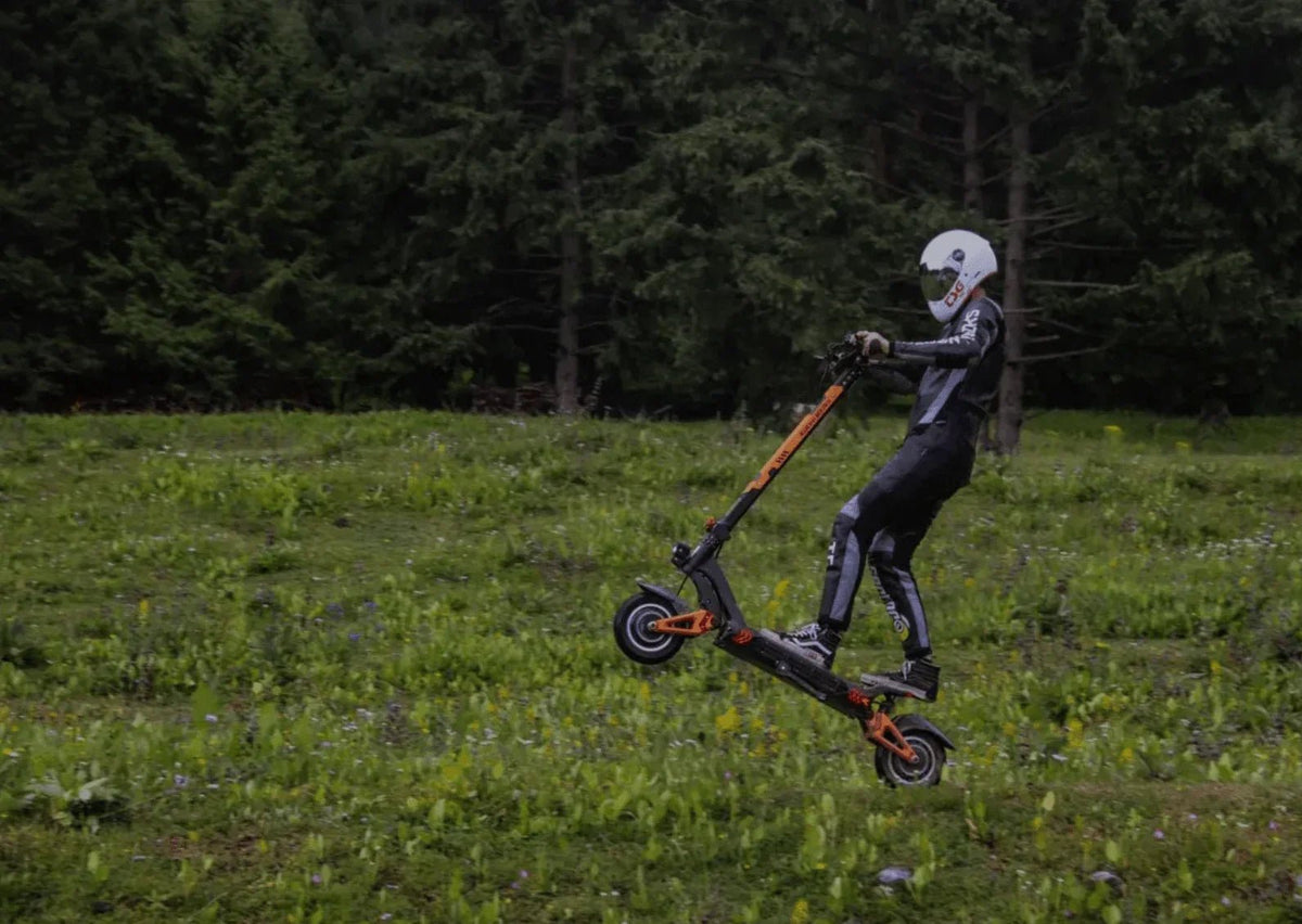 Fahrer mit weißem Helm und Schutzanzug springt mit einem schwarz-orangen E-Scooter über eine Wiese vor einem Fichtenwald