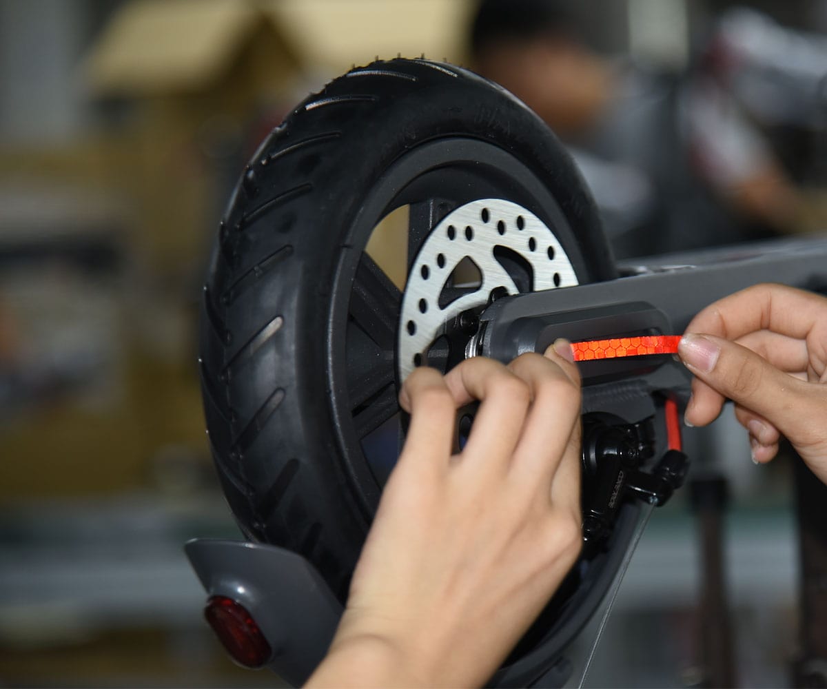 Hände kleben einen roten Reflektorstreifen an die Hinterradgabel eines E-Scooters, neben Profilreifen und Bremsscheibe