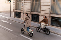 Zwei Personen fahren auf einer Stadtstraße jeweils auf einem Klapprad, eines beige und eines schwarz