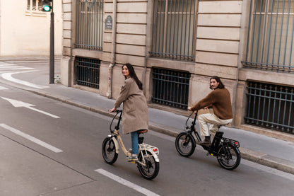 Zwei Personen fahren auf einer Stadtstraße jeweils auf einem Klapprad, eines beige und eines schwarz