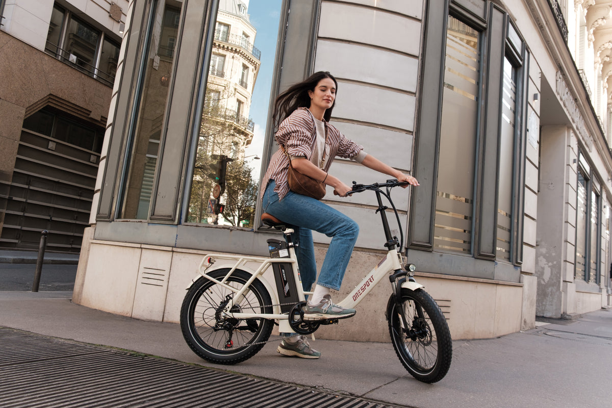 Frau fährt OneSport OT16 Pro, weißes E-Bike, Seitenansicht auf einer Stadtstraße vor einem Steingebäude