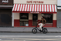 Mann fährt OneSport OT16 Pro, schwarzes Fahrrad, in Seitenansicht vor einem Café mit rot-weiß gestreifter Markise