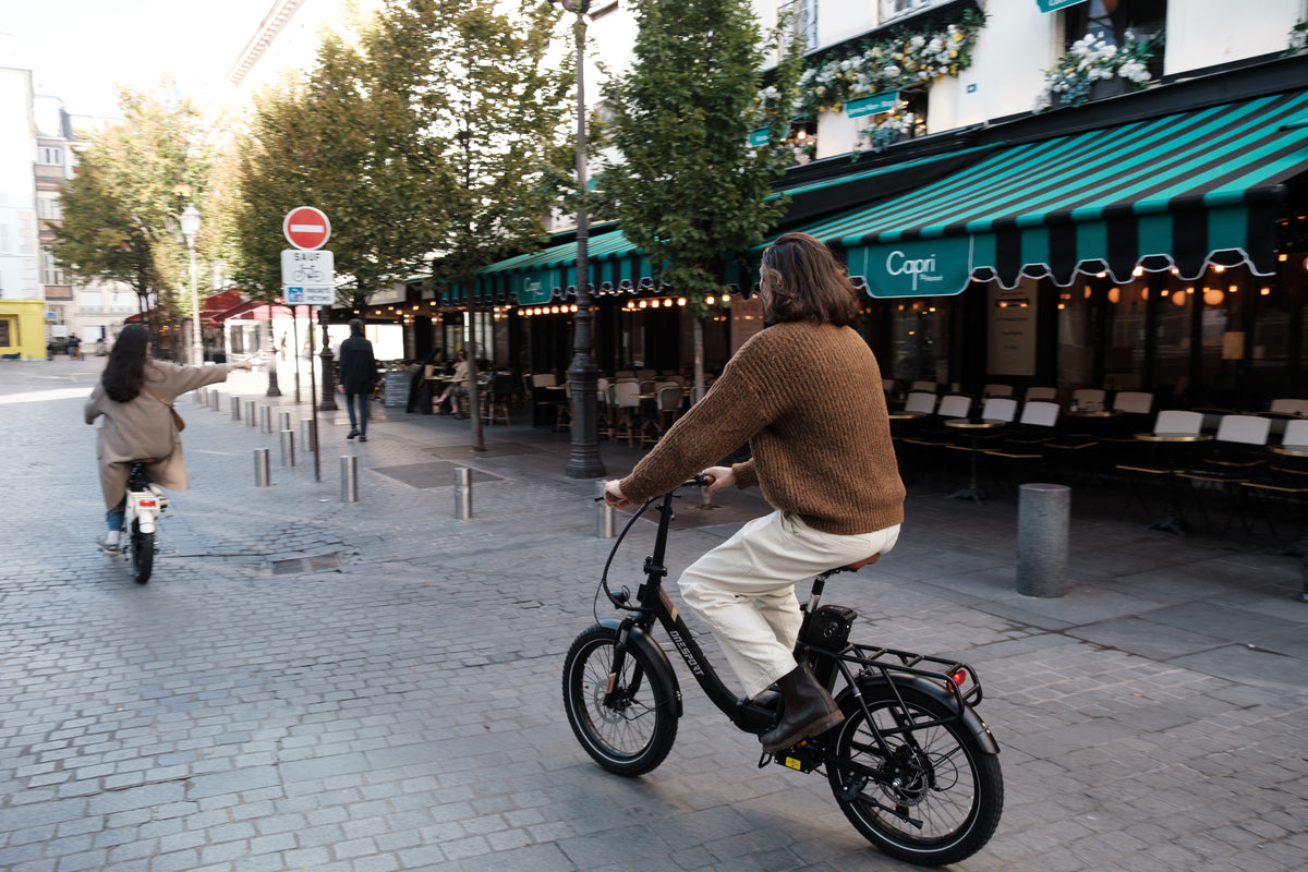 Person fährt OneSport OT16 Pro, schwarzes Fahrrad, auf einer Kopfsteinpflasterstraße an einem Café mit grün gestreifter Markise vorbei