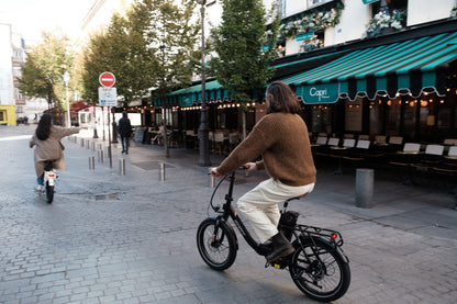 Person fährt OneSport OT16 Pro, schwarzes Fahrrad, auf einer Kopfsteinpflasterstraße an einem Café mit grün gestreifter Markise vorbei