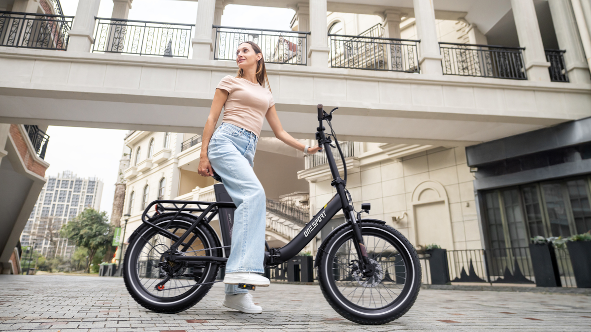 OneSport OT16 Pro: schwarzes Fahrrad in Seitenansicht mit einer Frau, die auf einem gepflasterten Platz daneben steht