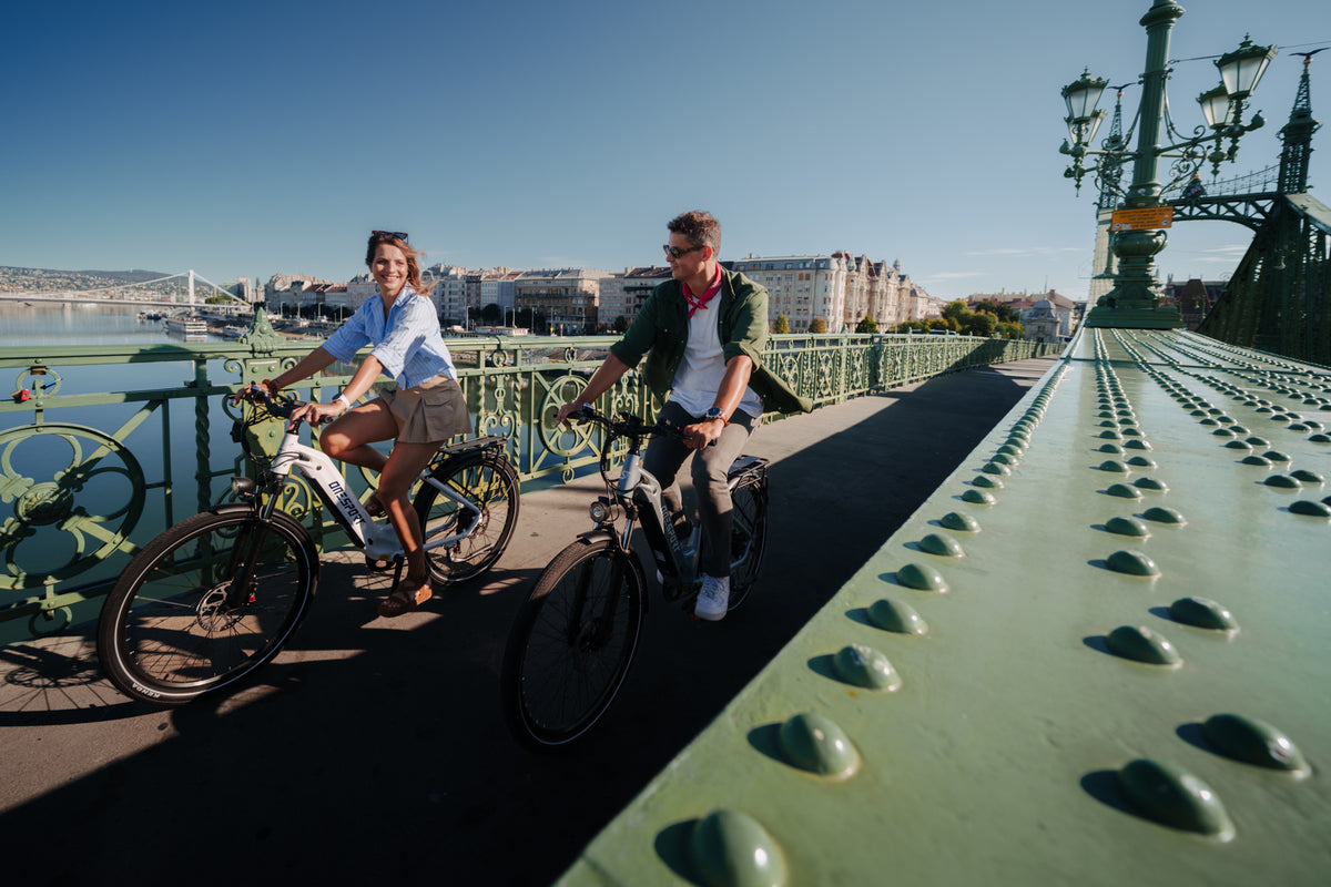 Zwei Personen fahren bei Sonnenschein auf einer grünen Brücke Fahrrad, im Hintergrund Gebäude und Wasser
