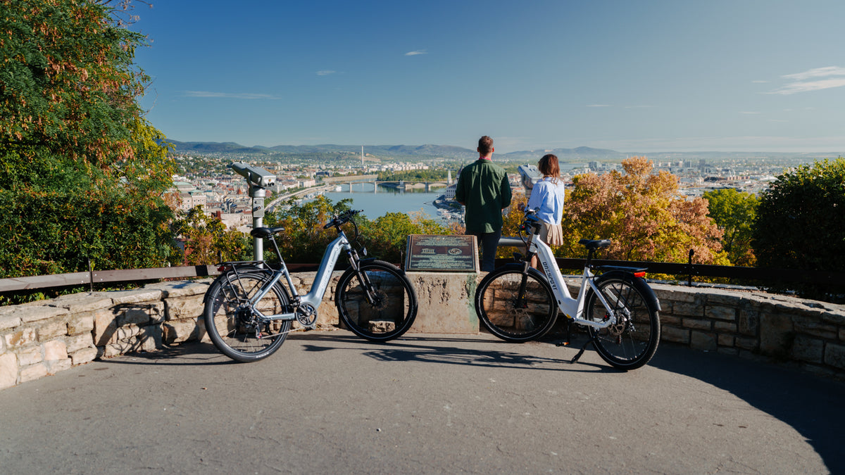 Zwei OneSport WT05 geparkt an einer Steinbrüstung an einem Aussichtspunkt, mit zwei Personen und Blick auf Stadt und Fluss