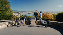 Zwei OneSport WT05 geparkt an einer Steinbrüstung an einem Aussichtspunkt, mit zwei Personen und Blick auf Stadt und Fluss