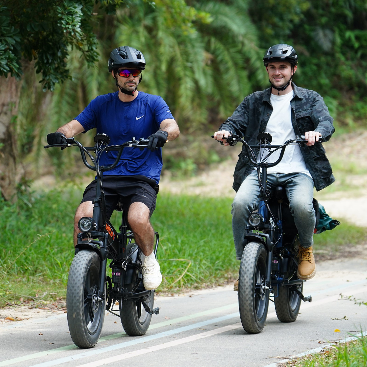 Zwei Personen mit Helm fahren nebeneinander auf Fatbikes über eine asphaltierte Straße im Grünen