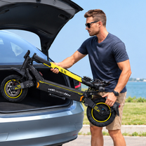 Mann mit Sonnenbrille hebt einen zusammengeklappten schwarz-gelben E-Scooter mit dem Schriftzug FREEBOY am offenen Kofferraum eines grauen Autos