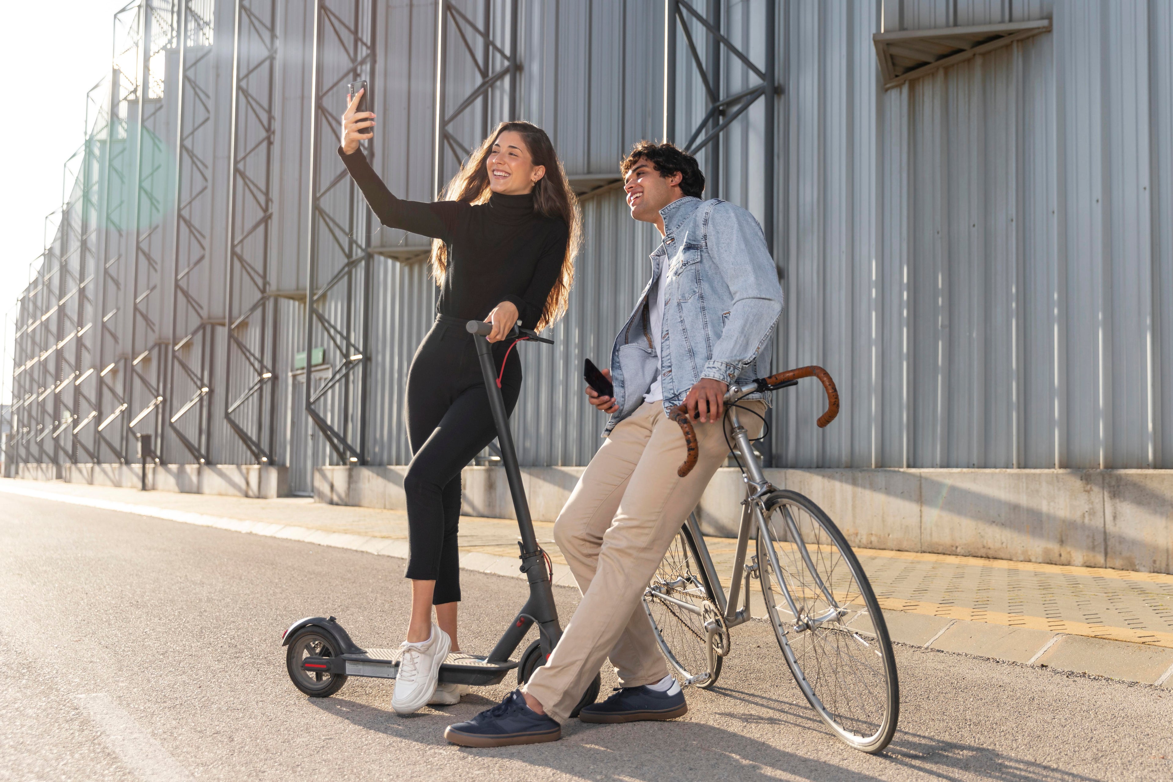 En man och en kvinna ståendes ute med en elsparkcykel och elcykel medan de tar en selfie med en smartphone