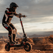 Person steht neben dem iScooter GX3 Pro E-Scooter auf sandigem Untergrund bei Sonnenuntergang